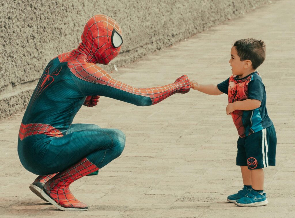 A child dressed in superhero attire shares a fist bump with a man in a Spiderman costume outdoors.